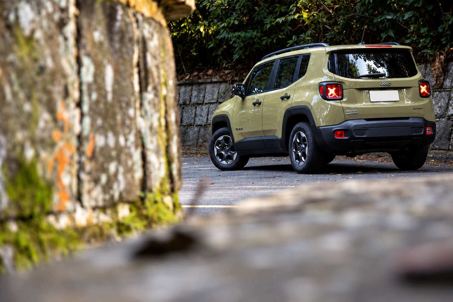 Jeep Renegade Sport 2.0 diesel 4x4 2017 verde-oliva, visto de três quartos traseiro, parado em uma estrada de serra asfaltada ao lado de um muro de pedras coberto de musgo, com vegetação densa ao fundo e luz suave de fim de tarde; as lanternas traseiras em formato de X estão acesas