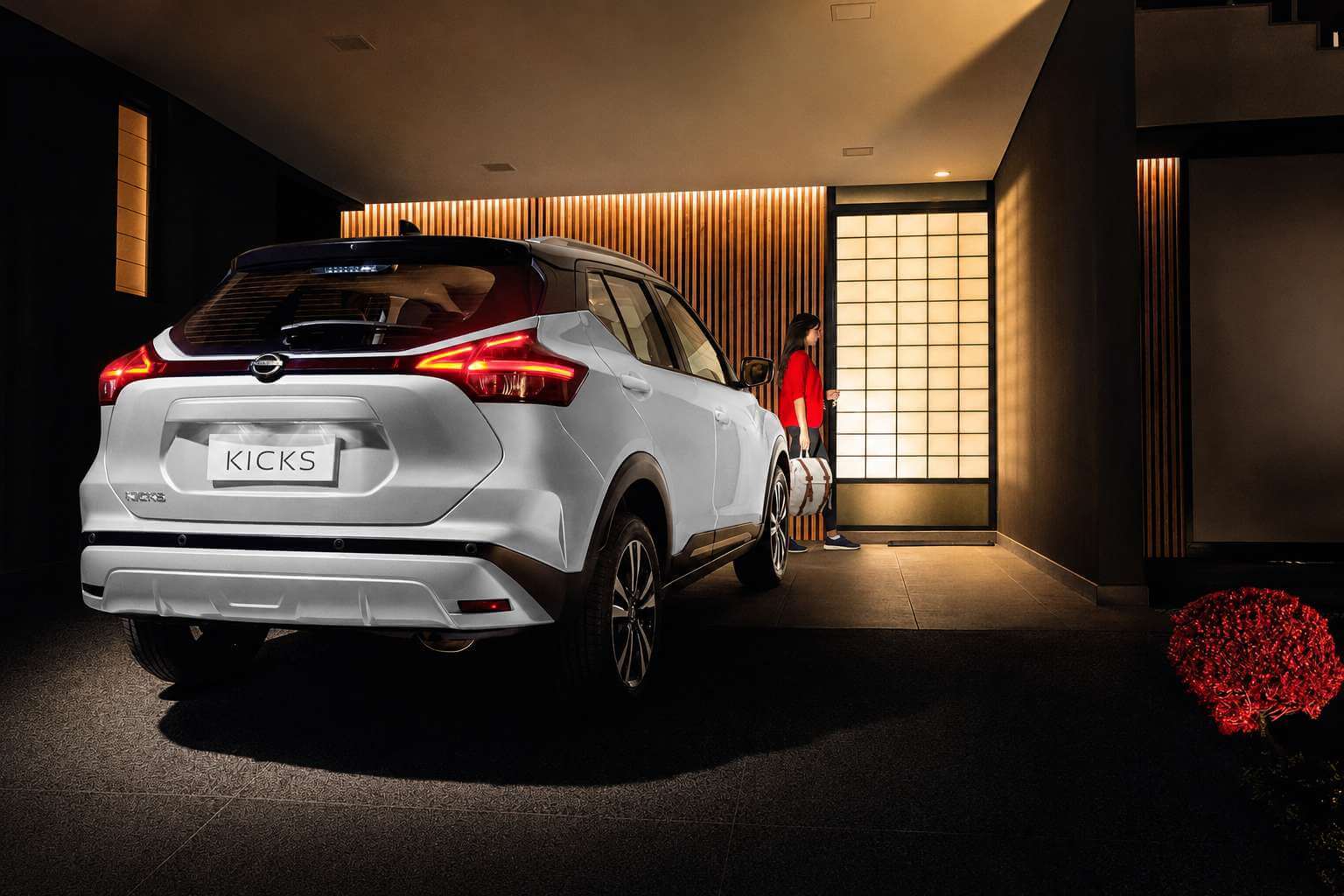 Nissan Kicks Advance branco de primeira geração, visto em três quartos traseiro dentro de uma garagem coberta de estilo japonês, com painéis de madeira ripada e porta de correr iluminada em luz quente ao fundo; aparecem as lanternas traseiras afiladas, o emblema Nissan e a inscrição Kicks na tampa, a placa com o nome Kicks e as rodas de liga diamantadas.