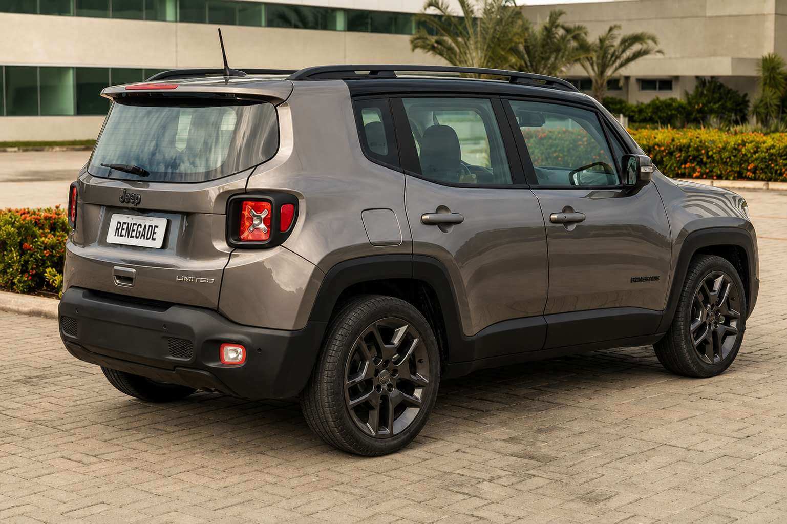 Jeep Renegade cinza fotografado de traseira em três quartos, parado num pátio de piso intertravado, com prédio de vidro e vegetação ao fundo; aparecem as lanternas traseiras em formato de X, o emblema Jeep na tampa e as rodas de liga escuras.