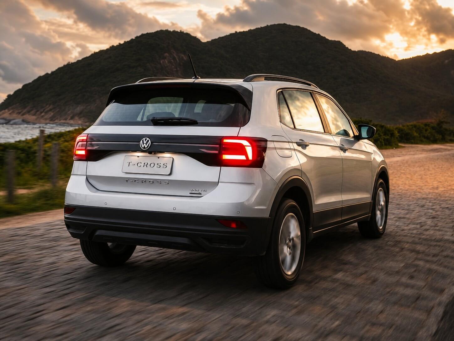 Volkswagen T-Cross branco visto em três quartos traseiro em movimento numa estrada litorânea ao pôr do sol, com morros e mar ao fundo; SUV compacto com lanternas de LED, o emblema da Volkswagen e a inscrição T-Cross na tampa, rodas de liga, com a inscrição T-Cross na placa.