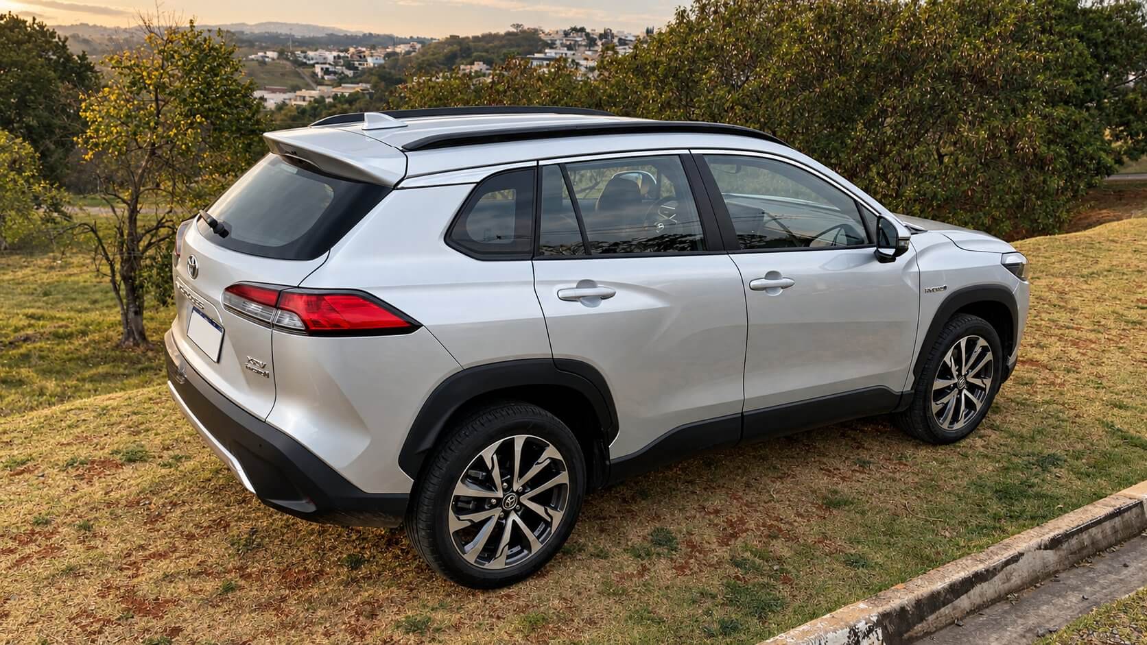 Toyota Corolla Cross Hybrid prata em três quartos traseiro, parado em um gramado com a cidade ao fundo