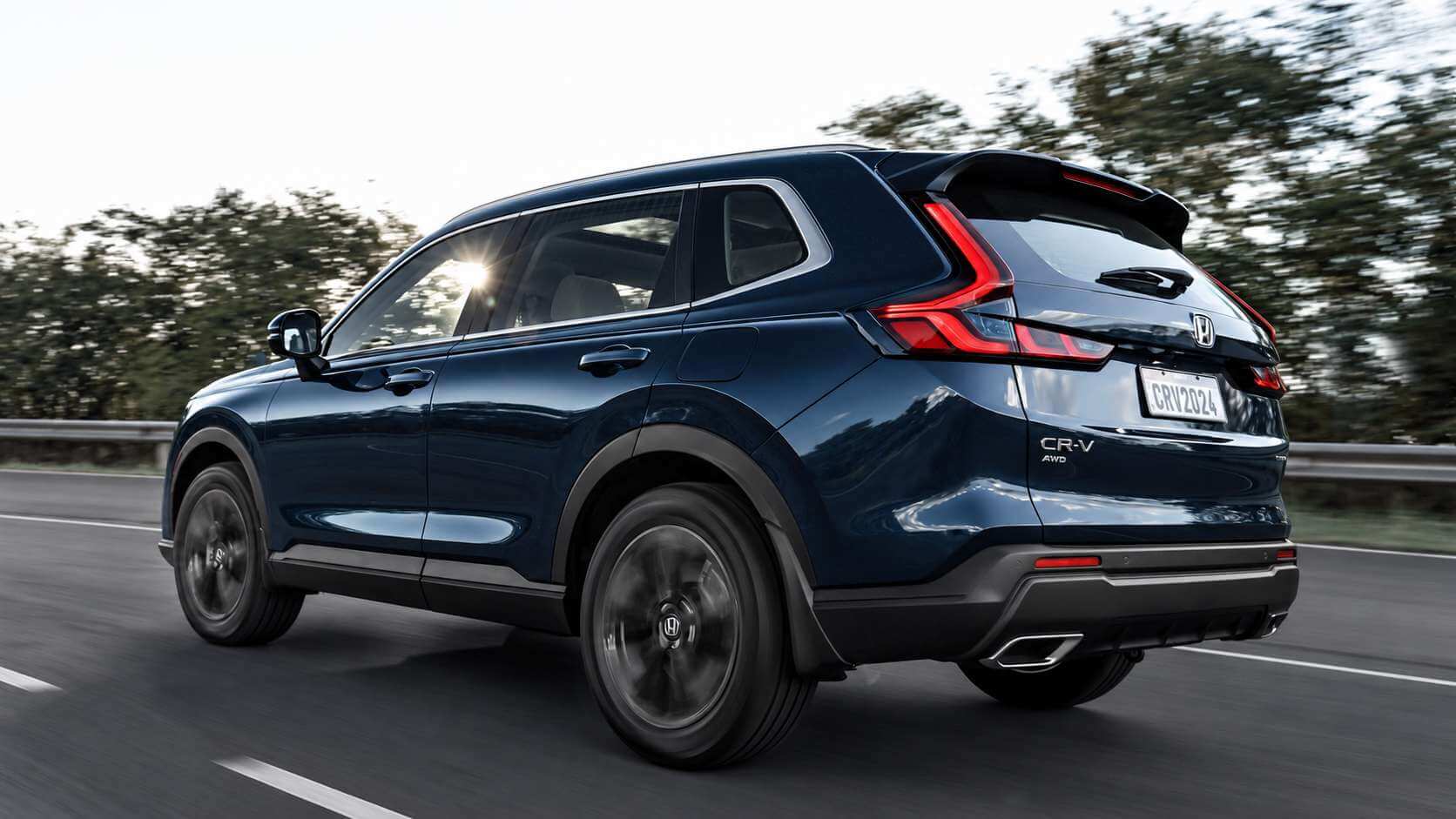 Honda CR-V Advanced e:HEV 2024 azul-escuro visto de tres quartos traseiro, em movimento em uma rodovia com arvores desfocadas ao fundo, sob luz de fim de tarde; aparecem o aerofolio de teto, as lanternas verticais em LED, o emblema Honda e o letreiro CR-V AWD na tampa traseira.