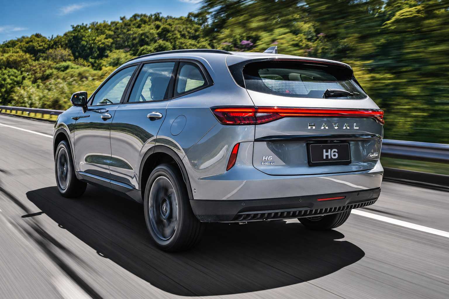 GWM Haval H6 1.5 turbo híbrido cinza visto em três quartos traseiro, rodando por uma rodovia com vegetação ao fundo