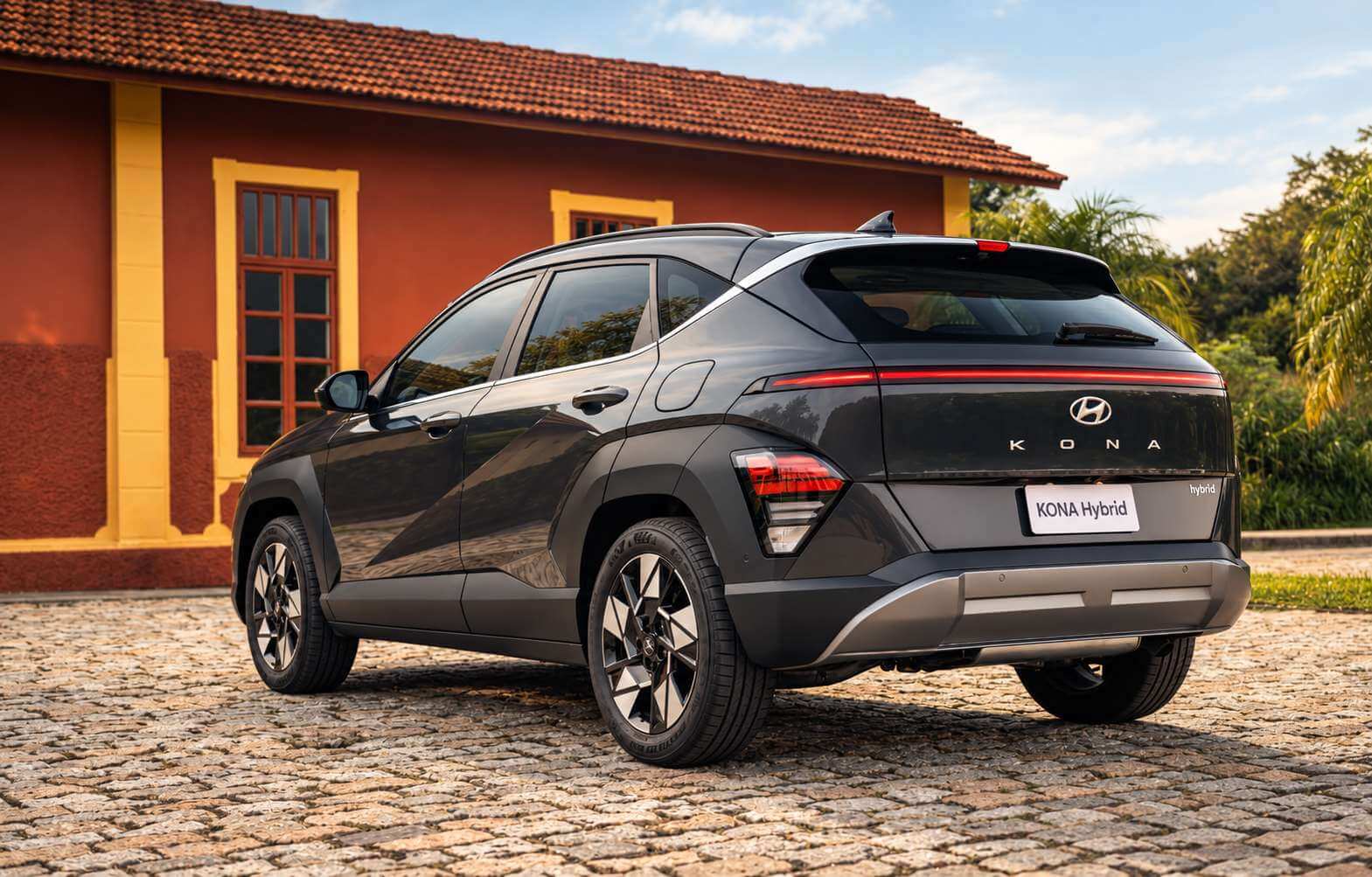 Hyundai Kona Hybrid cinza-escuro da segunda geração visto em três quartos traseiro, parado sobre um calçamento de pedras diante de uma casa colonial de paredes vermelhas e molduras amarelas, com palmeiras e mata ao fundo sob céu claro; a faixa de LED horizontal da tampa traseira e o letreiro KONA aparecem bem visíveis