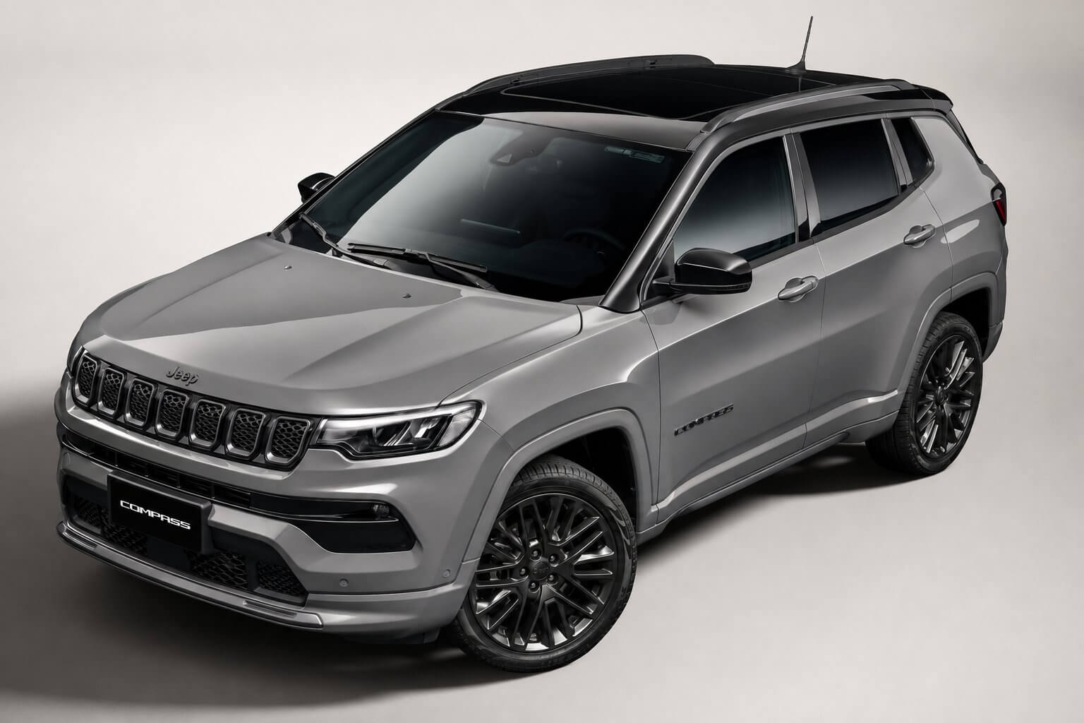 Jeep Compass cinza visto de cima em três quartos dianteiro, com o teto solar panorâmico de vidro em destaque no teto; SUV médio com o emblema da Jeep, a inscrição Compass na placa e rodas de liga escuras.