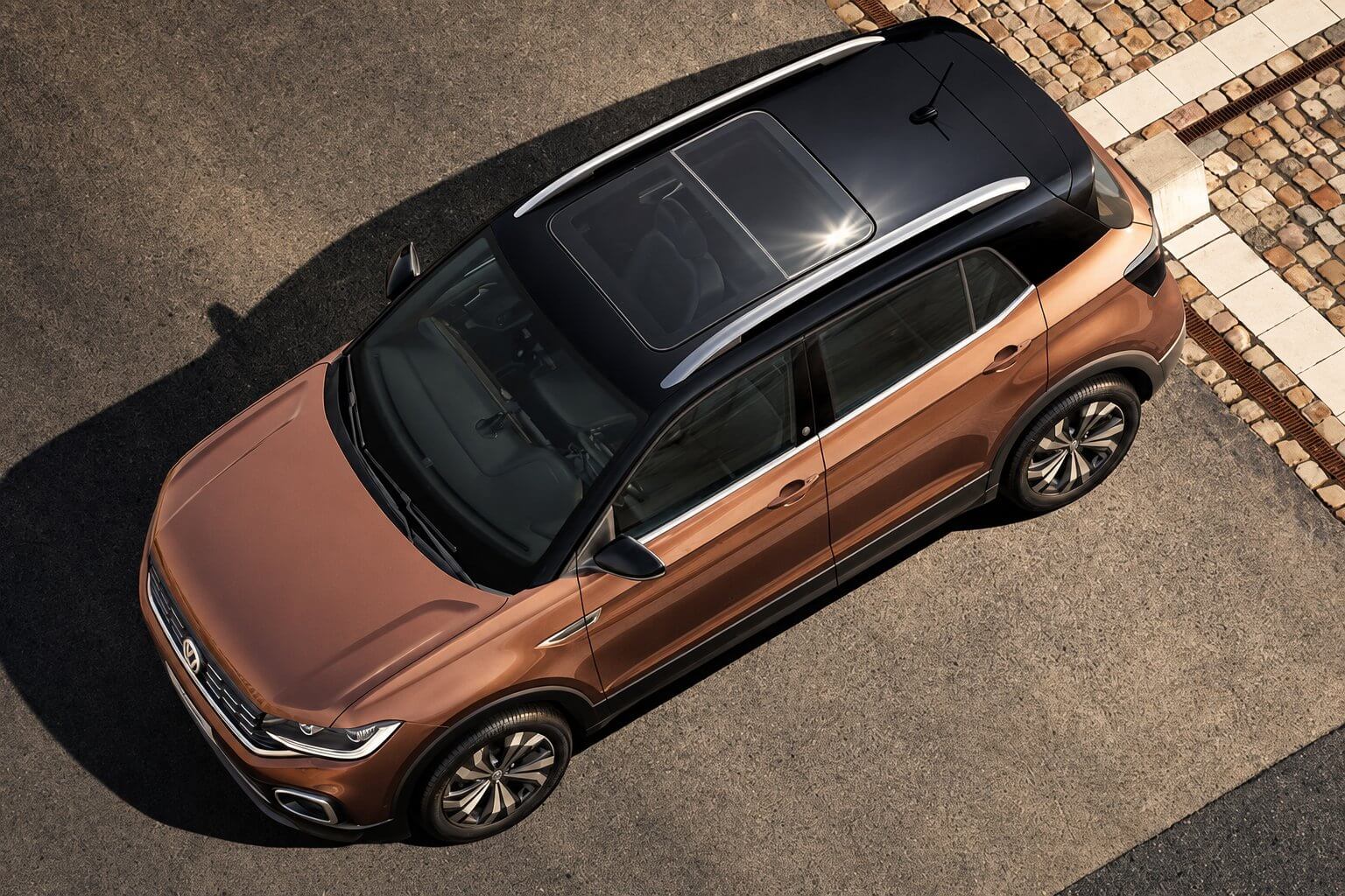 Volkswagen T-Cross marrom visto de cima, com o grande teto solar panorâmico de vidro em destaque no teto; SUV compacto com o emblema da Volkswagen, rack de teto e rodas de liga.