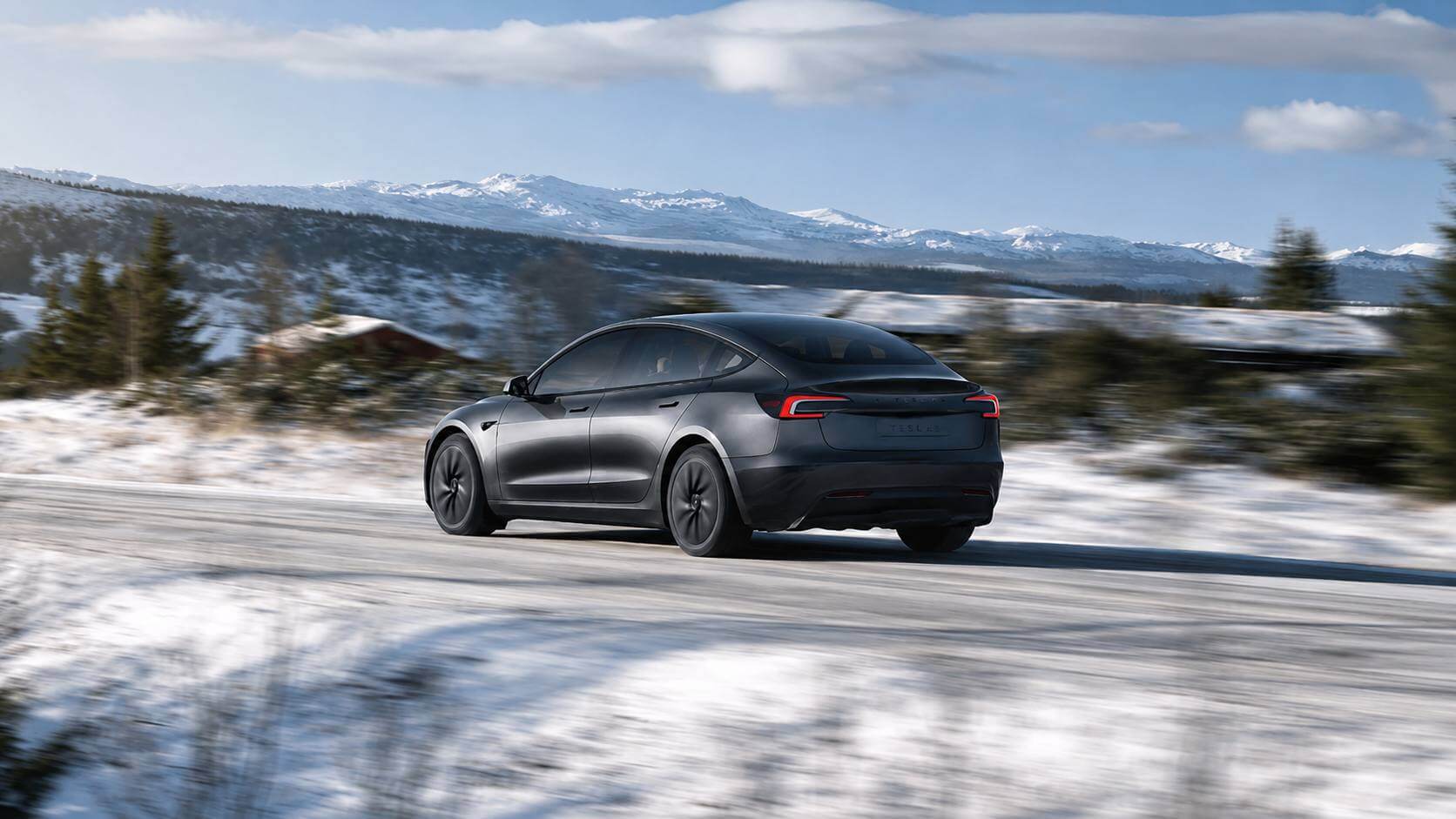Um Tesla Model 3 cinza visto de trás em três-quartos, em movimento numa estrada de montanha com neve e picos nevados ao fundo. Wordmark TESLA na tampa traseira.