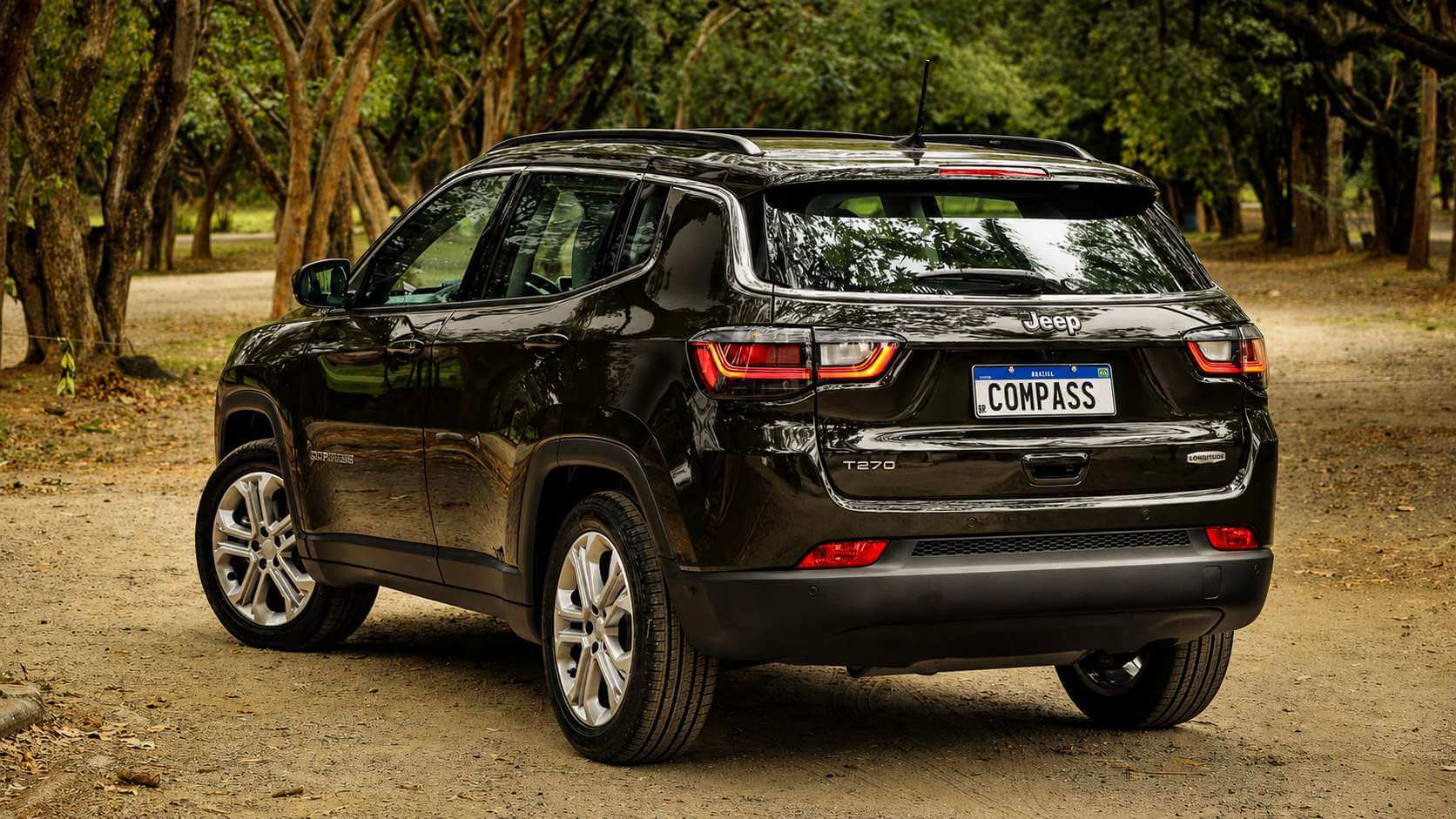 Jeep Compass Longitude T270 usado, de cor verde-escura quase preta, visto de trás em ângulo três quartos, parado em uma estrada de terra cercada por árvores altas e vegetação verde em um dia claro; a placa traseira traz apenas a palavra COMPASS
