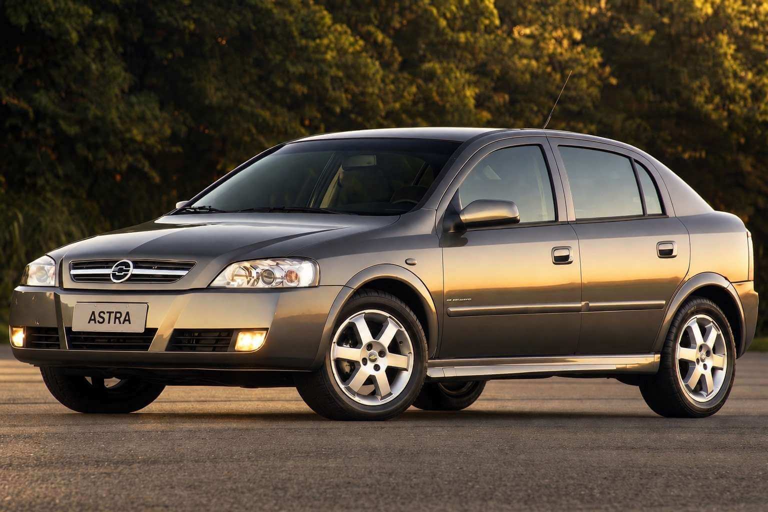 Chevrolet Astra hatch cinza-grafite de cinco portas visto em três quartos dianteiro, parado sobre o asfalto com árvores desfocadas ao fundo sob a luz dourada do fim de tarde; faróis de neblina acesos, rodas de liga leve de cinco raios e a placa dianteira em branco com a palavra ASTRA