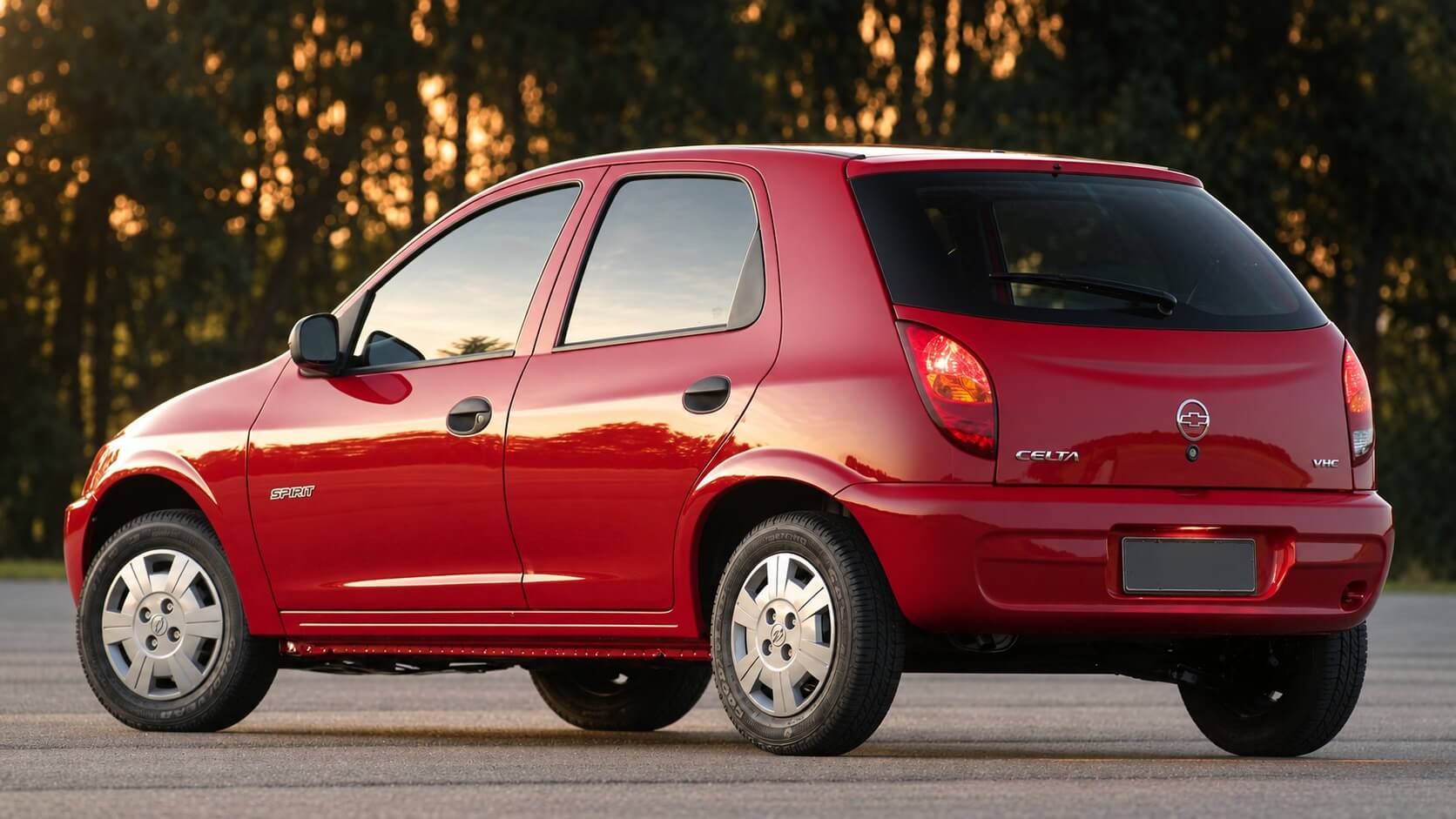 Chevrolet Celta hatch vermelho de cinco portas, visto em três quartos traseiro, parado sobre o asfalto de uma via larga com mata desfocada ao fundo, sob a luz dourada do fim de tarde; na tampa traseira aparecem o letreiro "CELTA", o emblema Chevrolet e a inscrição "VHC", com a placa em branco no para-choque, e na porta dianteira o emblema "SPIRIT"; calotas prateadas e faixa lateral escura completam a lateral do carro