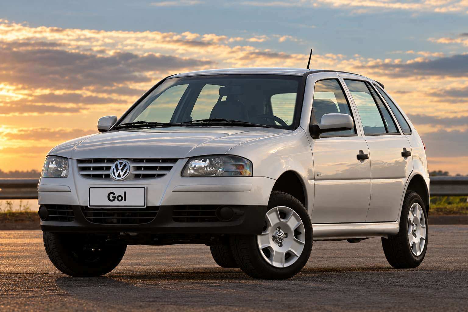 Volkswagen Gol G4 prata de quatro portas visto em três quartos dianteiro, parado sobre o asfalto de um acostamento com guard-rail e campo ao fundo, sob a luz alaranjada do fim de tarde; a grade dianteira traz o emblema da Volkswagen entre os faróis, as rodas são de liga leve de cinco raios e a placa exibe apenas a palavra Gol