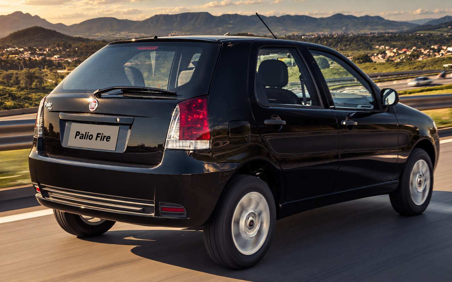 Fiat Palio Fire preto de cinco portas visto em três quartos traseiro, rodando por uma rodovia de pista dupla ao entardecer, com serras verdes e uma cidade ao fundo sob luz dourada; a lanterna vertical alta, o emblema da Fiat e o escrito Palio Fire na tampa do porta-malas aparecem nítidos