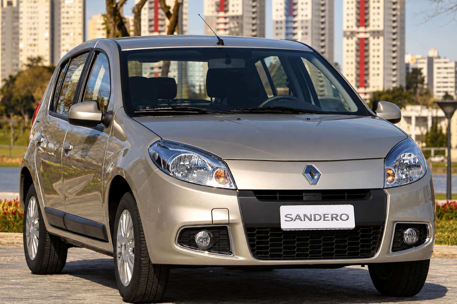 Renault Sandero da primeira geração reestilizada, na cor bege champanhe, visto em três quartos dianteiro, parado sobre um piso de pedra em uma área ajardinada; a grade preta traz o losango cromado da Renault ao centro, os faróis alongados avançam sobre os para-lamas e a placa dianteira mostra apenas a palavra SANDERO; ao fundo, prédios altos de uma cidade, árvores e canteiros de flores vermelhas sob luz natural de dia claro.
