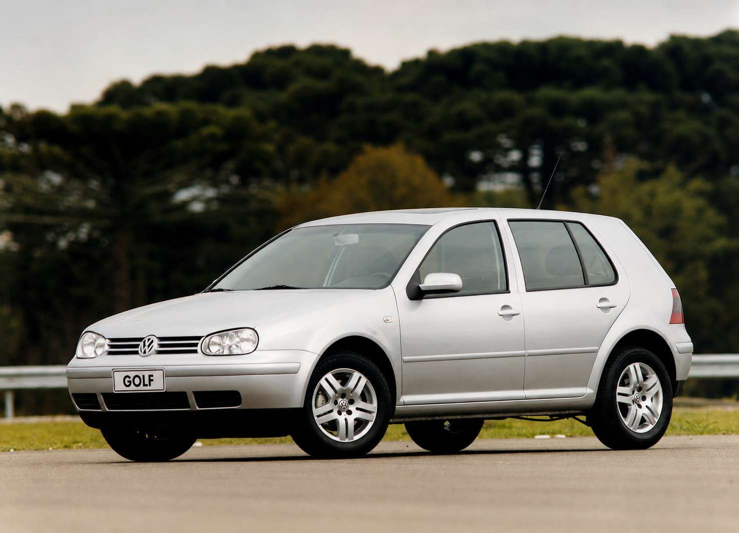 Volkswagen Golf GTI 1.8 Turbo 2004 na cor prata, visto em tres quartos dianteiro, parado no acostamento de uma estrada de asfalto; ao fundo, guard-rail metalico e uma mata fechada de araucarias sob ceu claro. O hatch de quatro portas traz farois duplos redondos, grade fina com o emblema da Volkswagen, retrovisores na cor da carroceria, rodas de liga leve de cinco raios duplos e placa dianteira com a inscricao GOLF.