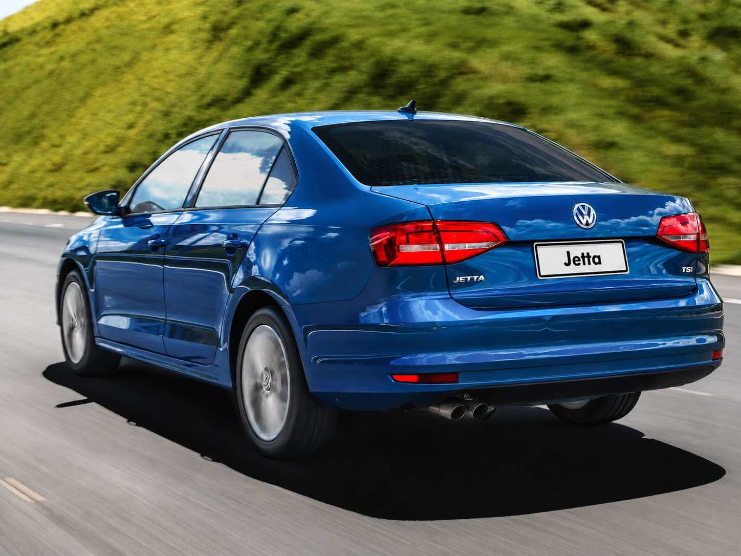 Volkswagen Jetta Comfortline 1.4 TSI 2016 na cor azul metálico, fotografado de trás em ângulo três quartos enquanto roda por uma estrada asfaltada, com uma encosta de vegetação verde ao fundo. Aparecem as lanternas traseiras horizontais avermelhadas, o emblema da Volkswagen e o letreiro Jetta na tampa do porta-malas, a placa com o nome do modelo, o emblema TSI no canto direito e as duas saídas cromadas do escapamento.
