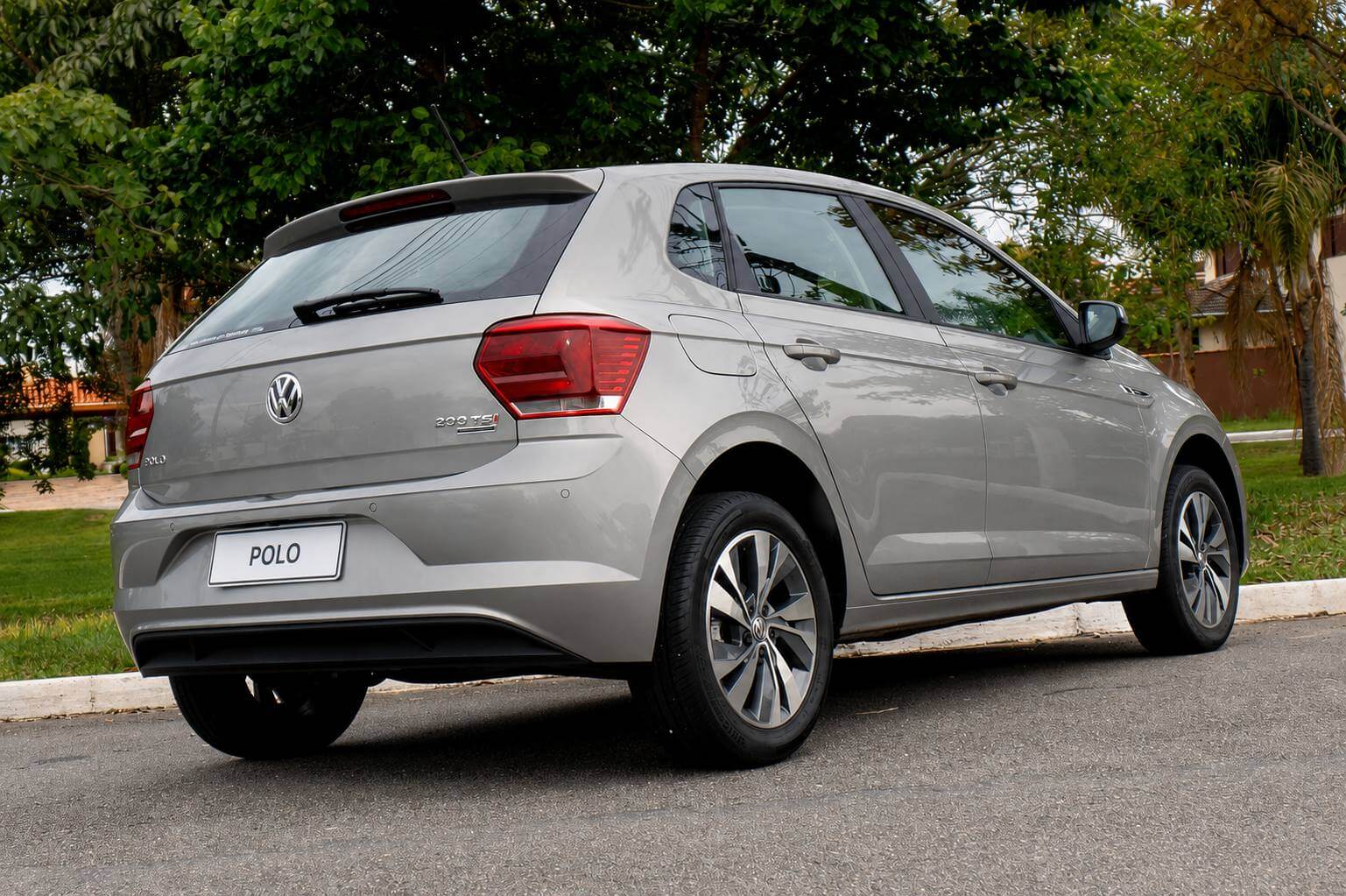 Volkswagen Polo Highline 1.0 200 TSI 2019 na cor prata, fotografado em tres quartos traseiro pelo lado direito, parado no asfalto de uma rua arborizada com gramado, meio-fio branco e casas ao fundo. Da para ver a lanterna vermelha vertical, o emblema da Volkswagen e a inscricao 200 TSI na tampa do porta-malas, o aerofolio sobre o vidro traseiro, a antena no teto, os sensores de estacionamento no para-choque e as rodas de liga leve de cinco raios duplos. A placa esta em branco, com apenas a palavra POLO.
