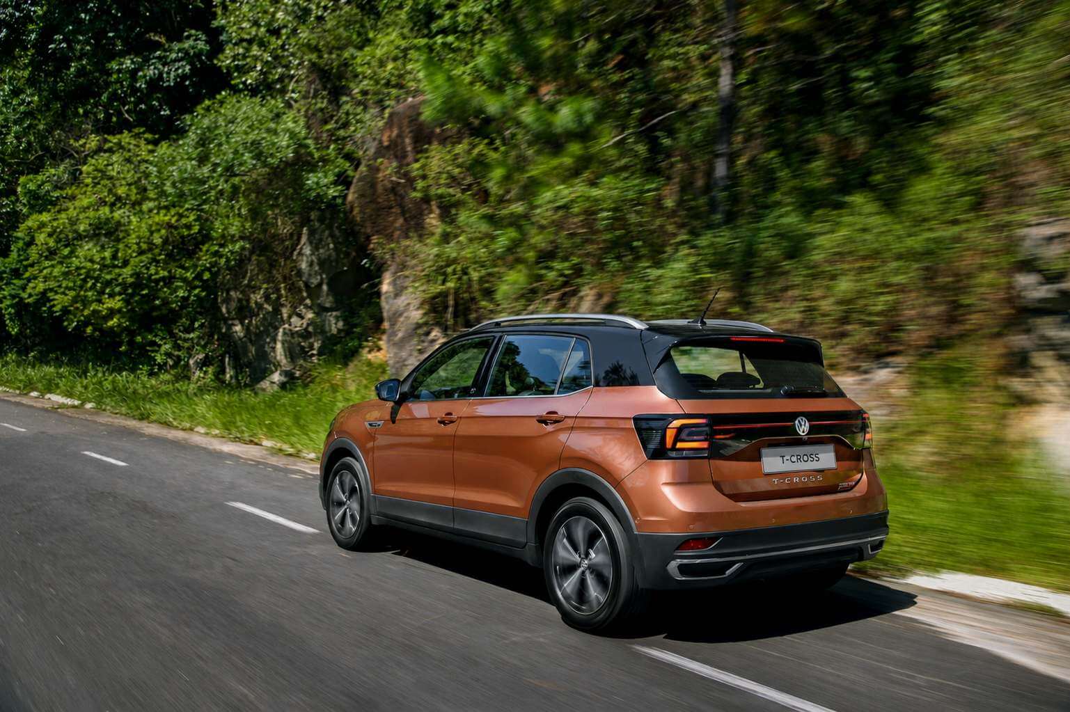 Volkswagen T-Cross Highline 250 TSI 2020 na cor cobre metálico, fotografado em três quartos traseiro pelo lado esquerdo enquanto roda por uma estrada de asfalto ladeada por vegetação verde e um paredão de rocha, com o fundo desfocado pelo movimento; aparecem as lanternas escurecidas, o emblema da Volkswagen e o letreiro T-CROSS na tampa traseira, as barras longitudinais no teto preto e as rodas de liga leve aro 17 de raios largos.