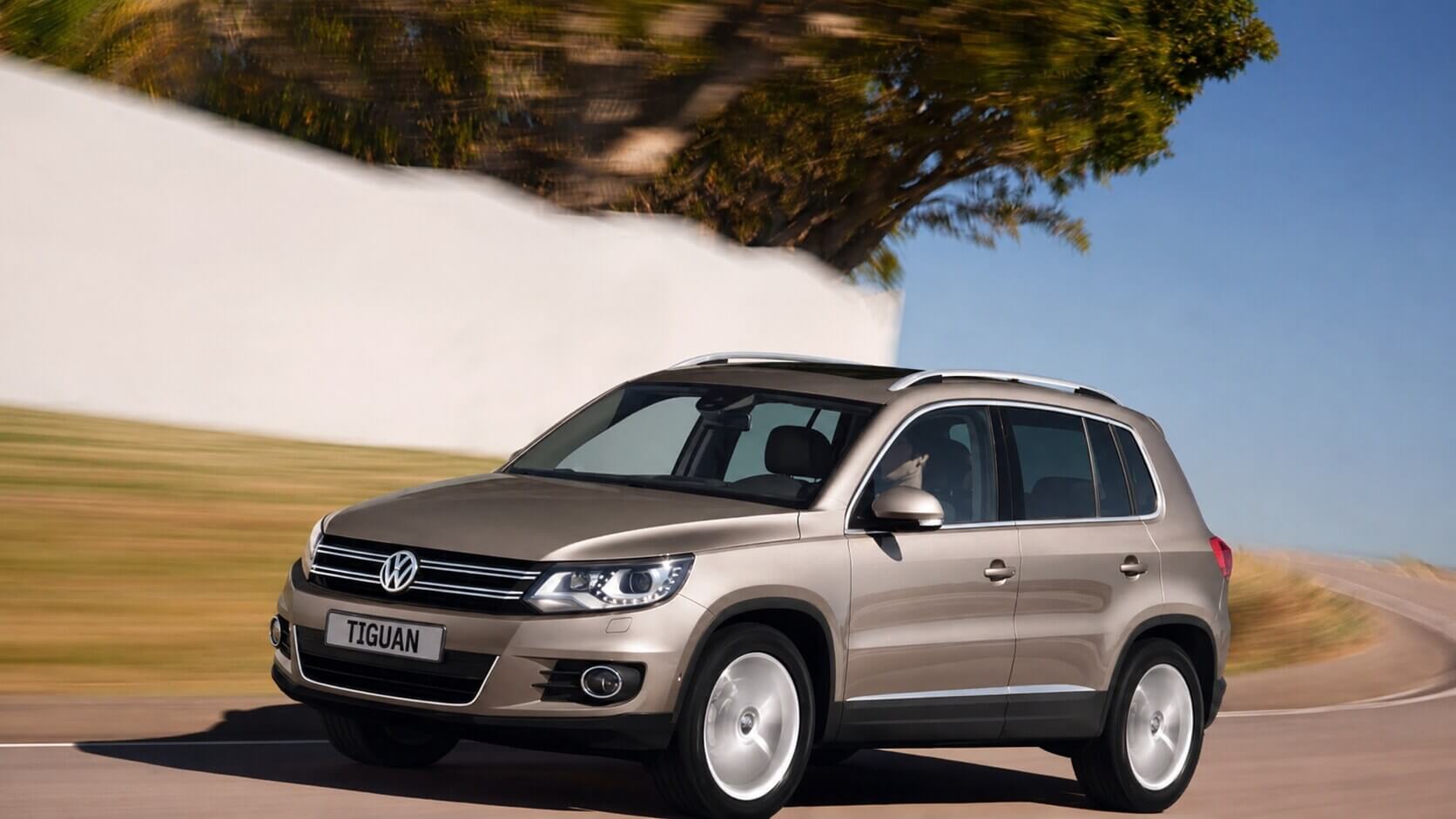 Volkswagen Tiguan 2.0 TSI 2012 na cor bege champanhe, fotografado em três quartos dianteiro enquanto roda por uma estrada de asfalto, com um muro branco, uma árvore de copa larga e um campo desfocado pelo movimento ao fundo. Aparecem a grade horizontal cromada com o emblema da Volkswagen, os faróis com luzes diurnas de LED, os faróis de neblina redondos no para-choque, as barras longitudinais prateadas no teto e as rodas de liga leve de cinco raios duplos. A placa dianteira está em branco, com apenas a palavra TIGUAN.