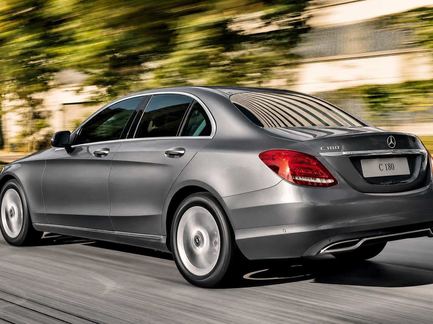 Mercedes-Benz C 180 cinza, geração 2016-2018 (W205), visto de traseira em três quartos rodando em uma via arborizada sob luz de fim de tarde; lanternas de LED acesas e a estrela de três pontas com a inscrição C 180 na tampa do porta-malas.