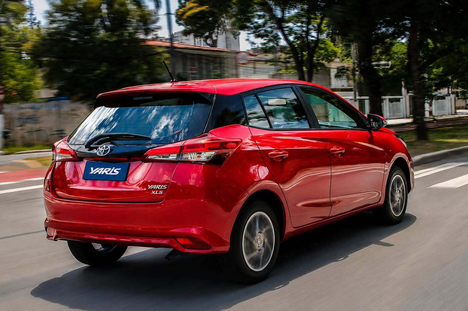 Toyota Yaris hatch vermelho fotografado em três quartos traseiro, em movimento por uma rua arborizada em dia claro; a tampa do porta-malas traz o emblema da Toyota, a placa com a inscrição Yaris e o emblema Yaris XLS, com lanternas afiladas, limpador traseiro, antena tipo vareta e rodas de liga leve de múltiplos raios
