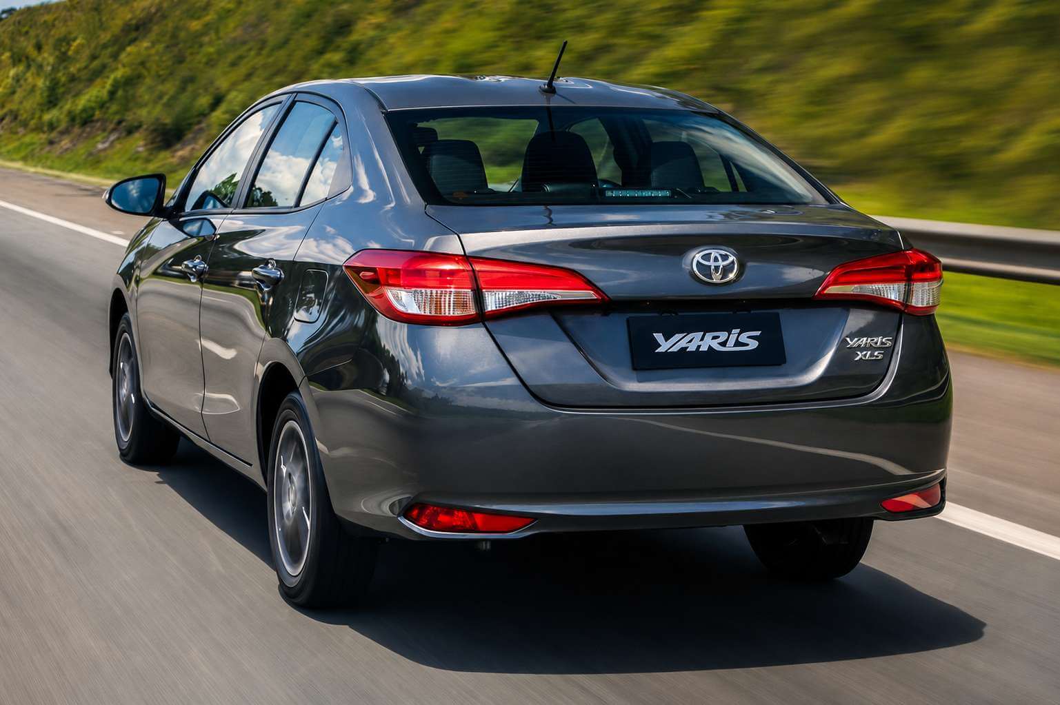 Toyota Yaris sedã na cor cinza-grafite fotografado em três quartos traseiro, em movimento por uma rodovia cercada de vegetação verde em dia ensolarado; a tampa do porta-malas traz o emblema da Toyota e a inscrição Yaris XLS, com lanternas vermelhas que se estendem em direção ao centro da traseira — a versão XL Plus Tech tem o mesmo desenho de carroceria, mudando basicamente as rodas
