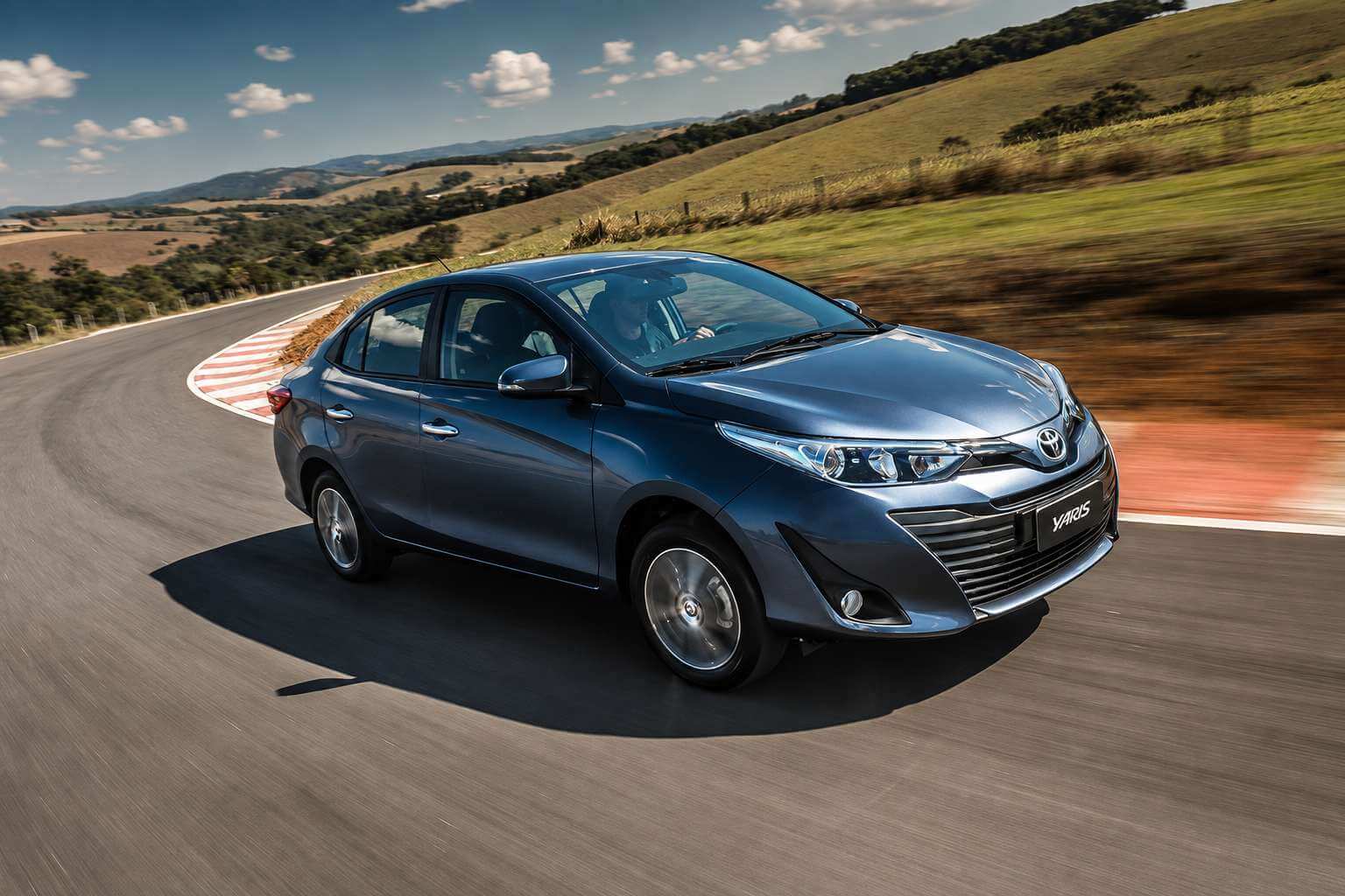 Toyota Yaris XLS Sedã 2020 na cor azul-grafite metálico, fotografado em três quartos dianteiro e em movimento numa curva de pista asfaltada, com colinas verdes, cerca de arame e céu azul com nuvens ao fundo em dia ensolarado; a grade dianteira em X traz o emblema da Toyota e a placa exibe apenas o nome Yaris, enquanto as rodas de liga leve aparecem com leve borrão de velocidade.