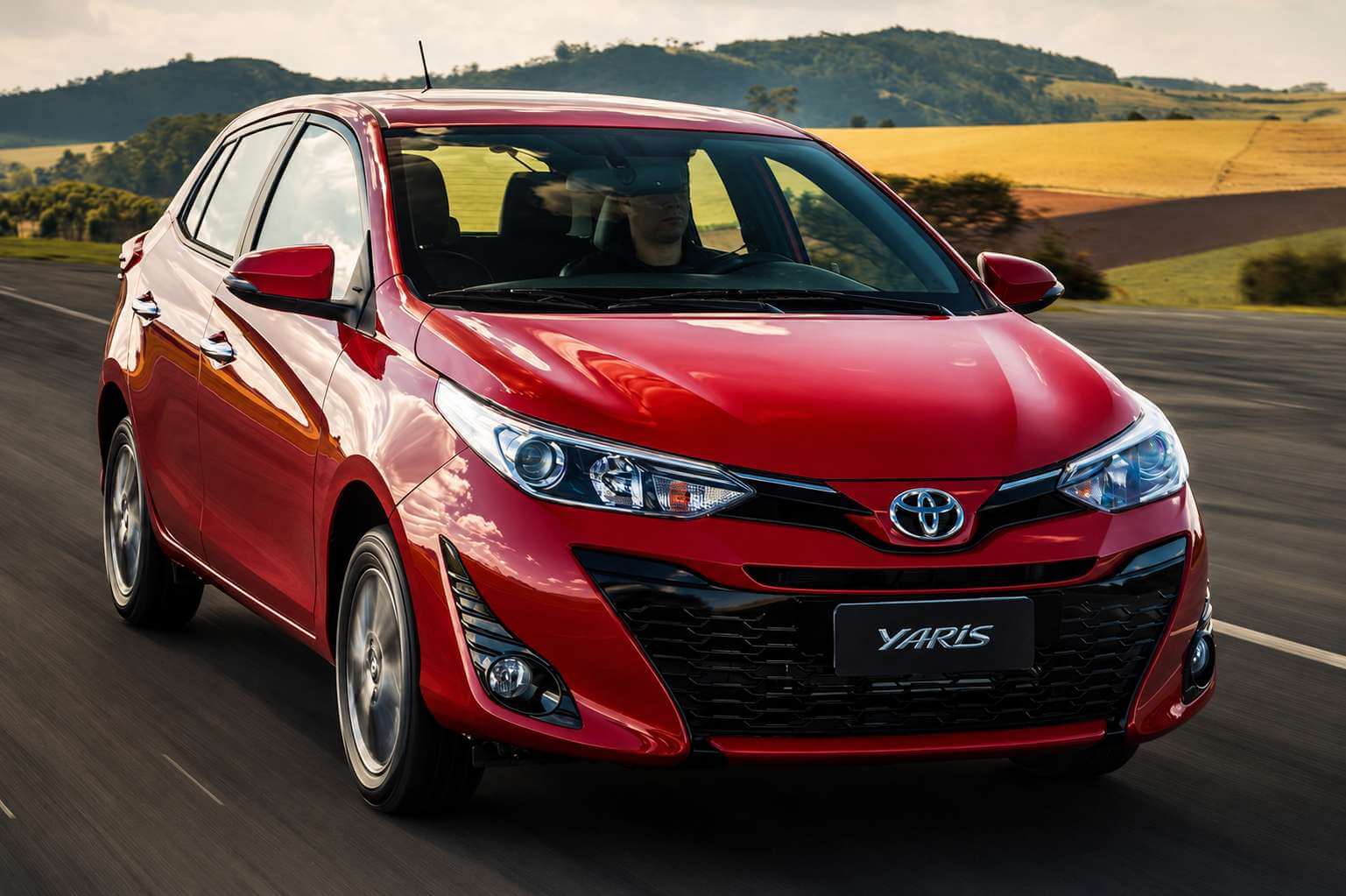Toyota Yaris hatch vermelho fotografado em três quartos dianteiro, em movimento por uma estrada de pista dupla no fim da tarde, com morros verdes e uma lavoura dourada ao fundo; a dianteira traz o emblema da Toyota no centro da grade, faróis com projetor, entradas de ar em X no para-choque, faróis de neblina, placa com a inscrição Yaris e rodas de liga leve de múltiplos raios
